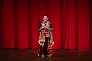 Woman in colorful traditional attire stands center stage, holding a microphone and smiling against red theater curtains