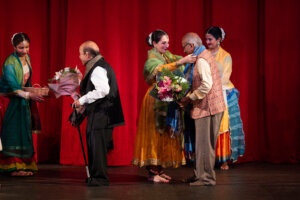 Group of performers in colorful Indian attire on stage, exchanging bouquets against a red curtain backdrop.