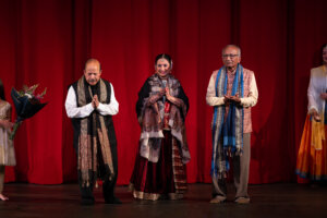 Group of performers in traditional Indian attire on a stage with a red curtain backdrop; center woman holds a microphone and smiles, flanked by two men in patterned shawls.