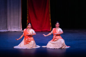 Two Indian classical dancers in orange and white costumes perform on stage with red and white backdrop curtains behind them, mid-dance pose and graceful hand gestures.