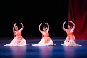Three female Bharatanatyam dancers kneel on a dark stage with orange and white costumes, arms raised in a dance pose, red curtain backdrop.