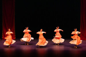Group of female Indian classical dancers in orange outfits performing on stage with red curtains in the background, mid-dance pose.
