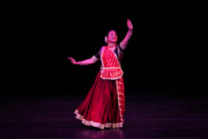 Female Indian classical dancer in a red and gold outfit on a dark stage, right hand raised in a graceful pose, smiling.