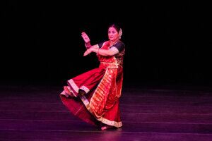 Female Indian classical dancer in a red and gold sari performing a pose on stage with a black background.
