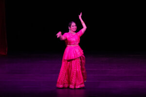 Female performer in a bright pink traditional dress dancing on a dark stage, arms raised and smiling.
