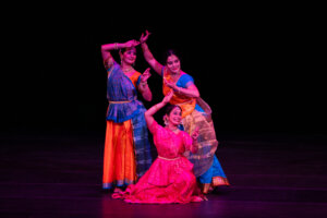 Group of female Indian classical dancers in bright sarees and jewelry performing on a dark stage backdrop. (Dance performance)