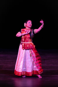 Female dancer in a red-and-gold traditional costume performing a classical pose on a dark stage with pink lighting