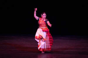Female dancer in a colorful red-and-gold sari performing a traditional Indian dance on a dark stage.