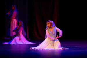 Dancer in a white and blue traditional costume with a blue veil kneels center stage, ornate jewelry, under purple stage lighting; two other performers in the background.