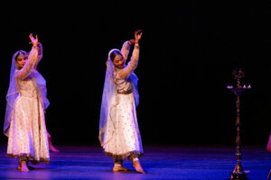 Three dancers in white outfits with blue veils perform on a dark stage, hands raised in graceful poses; a lit ceremonial stand with candles sits to the right.