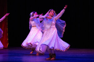 Dancers in pale blue, polka-dotted costumes performing a traditional dance on a dimly lit stage with purple lighting.