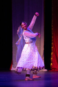 Female performer mid-dance on stage, wearing a white embroidered dress with a blue sheer veil and gold trim, arms raised in a graceful pose.