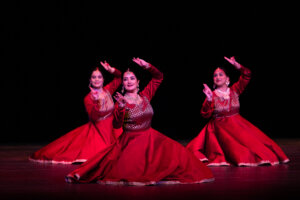 Three women in red traditional dresses perform a dance on a dark stage, one in the foreground with raised arms.