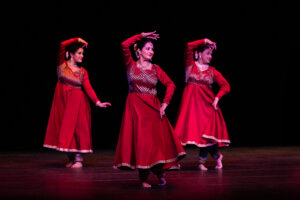 Three female dancers in red traditional costumes perform on a dark stage, mid-movement with elegant arm positions.