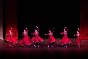 Group of female dancers in red traditional costumes perform a synchronized dance on a dark stage with a black backdrop.