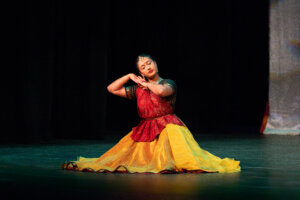 Female Indian classical dancer in a red and yellow sari, posed with hands folded at head level on a dark stage backdrop.