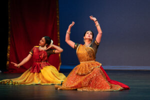 Two Indian classical dancers on stage, one in orange gold dress with raised arms, the other in red and yellow, performing a pose near a red curtain.