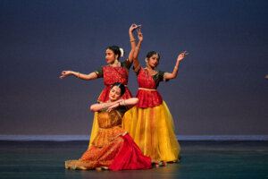 Three Indian classical dancers in red and gold costumes perform synchronized poses on a stage at dusk, wearing jewelry and flowers in their hair against a dark blue backdrop, with yellow skirts visible.