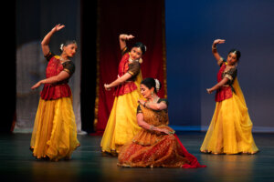 Group of female dancers in red and yellow traditional outfits performing onstage.