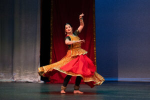 Indian classical dancer in a gold-and-orange traditional lehenga with jewelry, mid-mution pose on stage against a blue backdrop, arms raised and feet poised in a dance step.