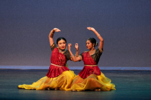 Two Indian classical dancers in red and yellow costumes performing a synchronized pose on a stage.
