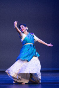 Indian classical dancer in a blue saree, mid-performance with raised arm and smiling on stage