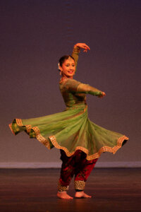 Indian classical dancer mid-performance in a green, gold-embroidered costume with arms raised in a graceful pose on stage, smiling.