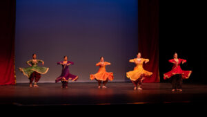 Five Indian classical dancers in colorful traditional outfits performing a synchronized spin on stage with red curtains.