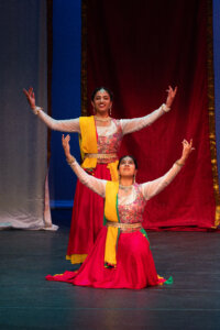 Two dancers in bright yellow and pink traditional outfits perform with arms raised on a stage.