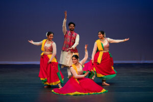 Group of Indian dancers in colorful traditional outfits performing on a stage with a dark blue backdrop; central male dancer raises his hand while others pose gracefully around him.