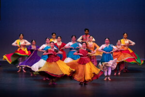 Group of performers in bright traditional Indian costumes dancing on a stage with a dark blue backdrop, colorful flowing skirts swirling.