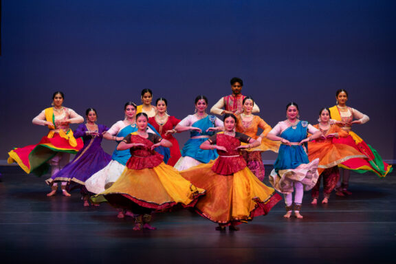 Group of performers in bright traditional Indian costumes dancing on a stage with a dark blue backdrop, colorful flowing skirts swirling.