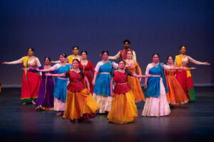 Group of Indian classical dancers in colorful costumes performing on stage with blue backdrop.