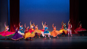 A group of colorful Indian dancers performing on stage in vibrant costumes, mid-movement with flowing skirts.