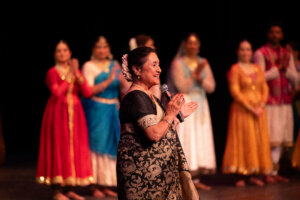 Smiling woman in a black and gold sari sings into a microphone on stage, with colorful dancers in festive outfits blurred in the background.