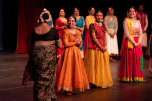 Group of Indian classical dancers on stage in bright sarees and lehengas, smiling after a performance, audience not visible.