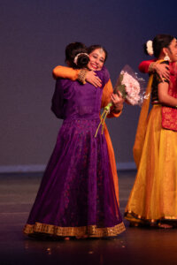 Two women in vibrant traditional dresses hug on stage; one holds a bouquet of pink flowers and smiles warmly as they embrace.