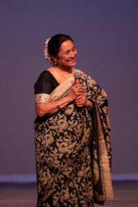 Elderly Indian woman in a black and gold saree, holding a microphone and smiling on stage with flowers in her hair.