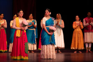 Group of Indian dance performers on stage in bright traditional outfits; two women in front hold bouquets and smile as others applaud behind them.