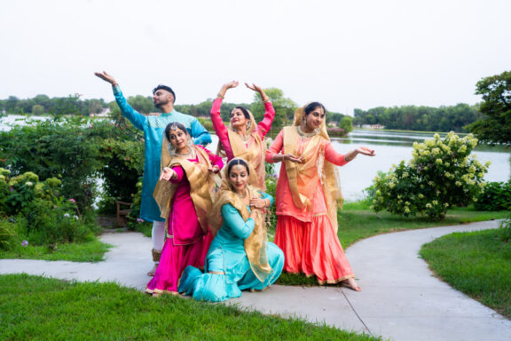 Group of people in bright traditional Indian clothing dancing outdoors by a lake and greenery.